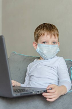Karantina altındaki çocuklar. Kendini soyutlama. Tıbbi maskeli bir çocuk kanepede otururken dizüstü bilgisayarda çizgi film izler. Çocuklar için yasak şeyler, aletler..