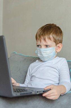 Karantina altındaki çocuklar. Kendini soyutlama. Tıbbi maskeli bir çocuk kanepede otururken dizüstü bilgisayarda çizgi film izler. Çocuklar için yasak şeyler, aletler..