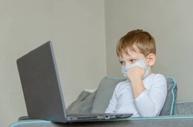 Karantina altındaki çocuklar. Kendini soyutlama. Tıbbi maskeli bir çocuk kanepede otururken dizüstü bilgisayarda çizgi film izler. Çocuklar için yasak şeyler, aletler..