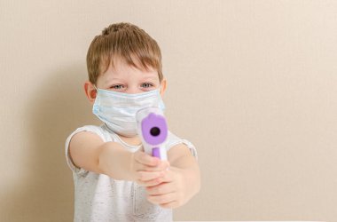 Tıbbi maske takan dört yaşında tatlı bir çocuk doktorculuk oynuyor ve sıcaklığı temassız termometreyle ölçüyor. Koronavirüs salgınından dolayı çocuklarla kendini izole etme