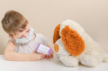 Tıp maskesi takmış dört yaşında tatlı bir çocuk doktorculuk oynuyor ve oyuncak peluş köpeğinin sıcaklığını temassız termometreyle ölçüyor. Koronavirüs salgınından dolayı çocuklarla kendini izole etme
