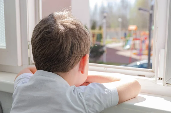 4歳の可愛い悲しい少年がバルコニーに座って遊び場で窓の外を見ています 世界のコロノウイルスのパンデミック による自己分離 — ストック写真
