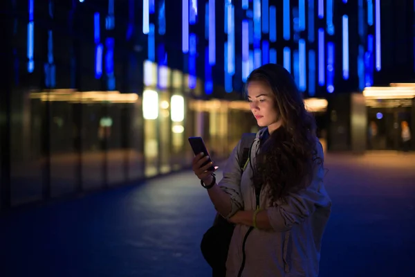 Mooie stijlvolle jongedame wandelen in de straten van de nacht voor de stad met behulp van moderne smartpone — Stockfoto