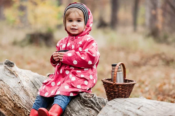 Маленька дівчинка в гороховій куртці сидить на дереві і п'є чай . — стокове фото