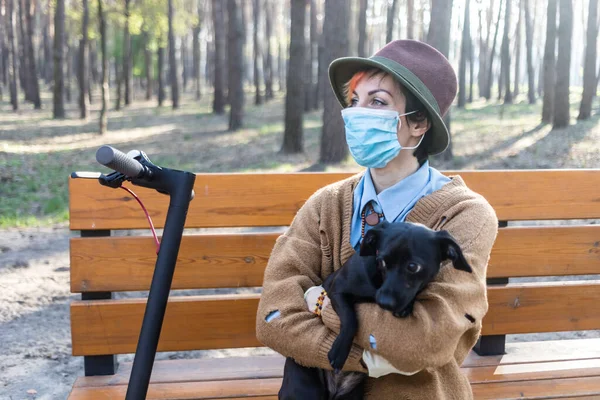 コロナウイルスから身を守るためにマスクと手袋をしたスタイリッシュな女性 市内や公園内の電動スクーターを使用して安全になります 犬と遊ぶのは楽しい — ストック写真