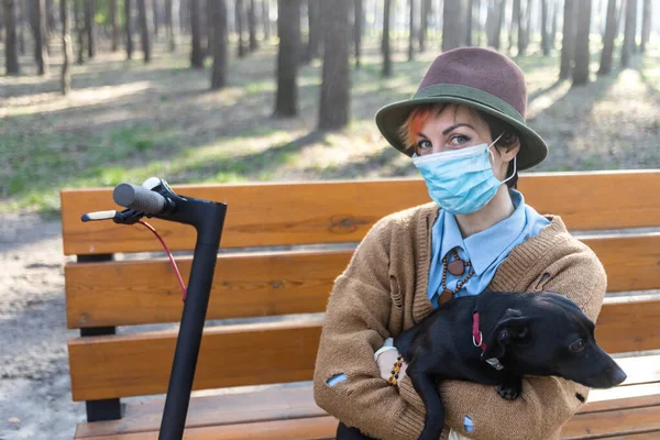 コロナウイルスから身を守るためにマスクと手袋をしたスタイリッシュな女性 市内や公園内の電動スクーターを使用して安全になります 犬と遊ぶのは楽しい — ストック写真