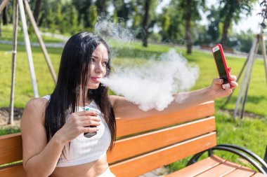 Çekici kadın sigara vape cihaz, parkta telefonda haber okuma.
