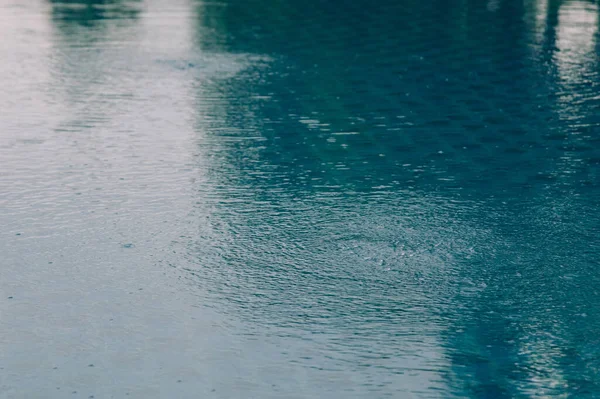 rain drops on the surface of water in a puddle with graduated shade of black shadow and reflection of blue sky