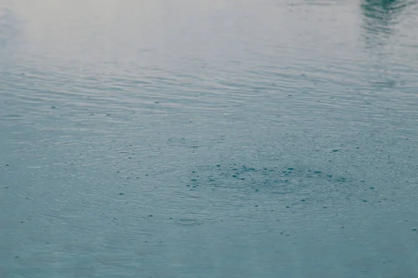 rain drops on the surface of water in a puddle with graduated shade of black shadow and reflection of blue sky