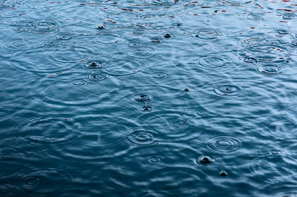 rain drops on the surface of water in a puddle with graduated shade of black shadow and reflection of blue sky