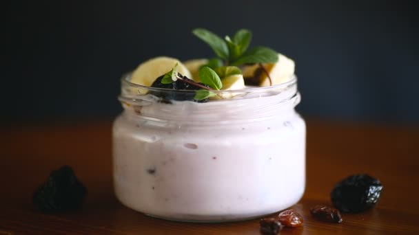 甜而好吃的酸奶 有香蕉 葡萄干 放在木制桌子上的玻璃瓶里 — 图库视频影像