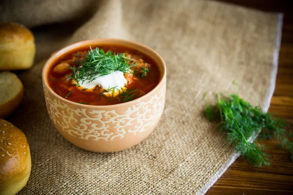 hot beetroot soup with sour cream, herbs and rolls in a ceramic bowl on a wooden table