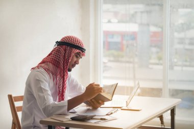 Arap Suudi adam online çalışma ile bir dizüstü bilgisayar ve kitap okuma 
