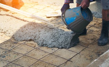 İnşaat işçisinin eldivenli ellerine yakın çekim için mala kullanarak ahşap formlar ve yeni dökülmüş beton bloknotlarla fazla karışım seviyesini kazıyın.