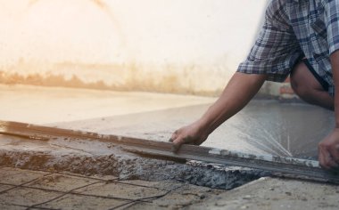 İnşaat işçisinin eldivenli ellerine yakın çekim için mala kullanarak ahşap formlar ve yeni dökülmüş beton bloknotlarla fazla karışım seviyesini kazıyın.