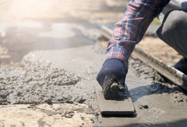İnşaat işçisinin eldivenli ellerine yakın çekim için mala kullanarak ahşap formlar ve yeni dökülmüş beton bloknotlarla fazla karışım seviyesini kazıyın.