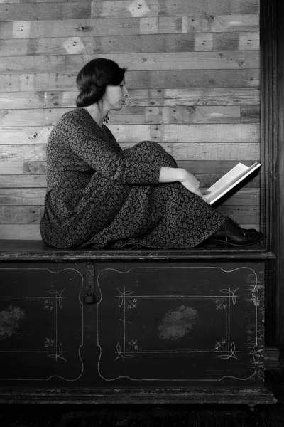 Mulher bonita senta-se em um peito preto e lê um livro sobre um fundo de madeira — Fotografia de Stock