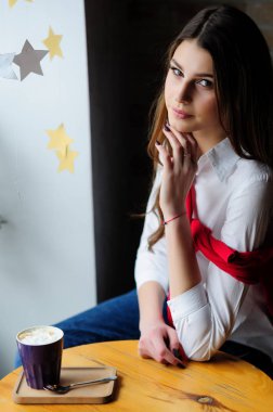 Bir kafede, pencerenin yanındaki masada bir fincan kahveyle oturan kız.