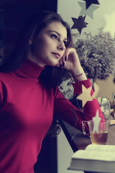 一个穿着红色毛衣的漂亮姑娘在一家咖啡馆的橱窗里 一杯茶和一本书 — 图库照片
