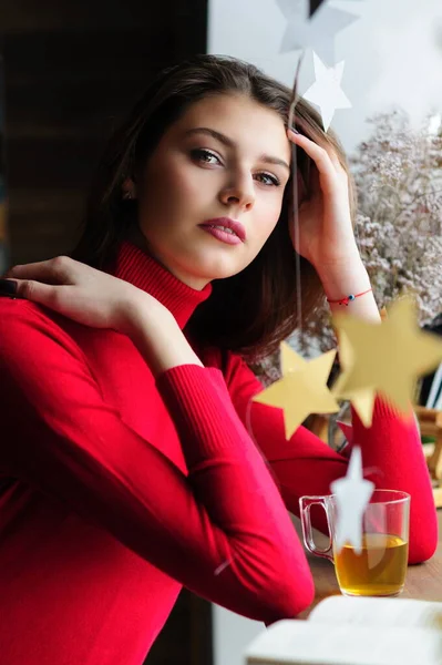お茶と本が入ったカフェの窓の赤いセーターを着た美しい女の子 — ストック写真