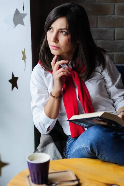 一个漂亮的黑发姑娘穿着白衬衫 坐在咖啡店的桌子边 边喝咖啡边看书的画像 — 图库照片