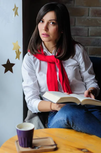 一个漂亮的黑发姑娘穿着白衬衫 坐在咖啡店的桌子边 边喝咖啡边看书的画像 — 图库照片