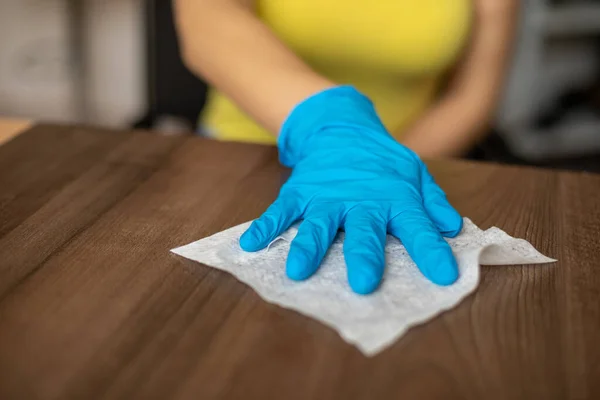 Woman\'s hand in blue gloves sanitizing cleaning home office wood table surface with wet wipes