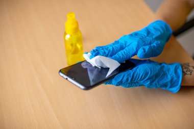 Mavi eldivenli bir kadın ahşap masa yüzeyinde ıslak mendil ve alkollü akıllı telefon temizliyor.