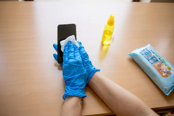 Woman\'s hand in blue gloves sanitizing cleaning smartphone mobile phone on wood table surface with wet wipes and alcohol