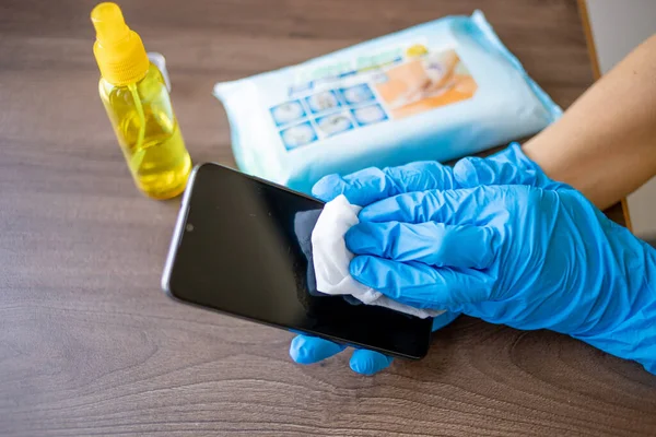 Woman\'s hand in blue gloves sanitizing cleaning smartphone mobile phone on wood table surface with wet wipes and alcohol