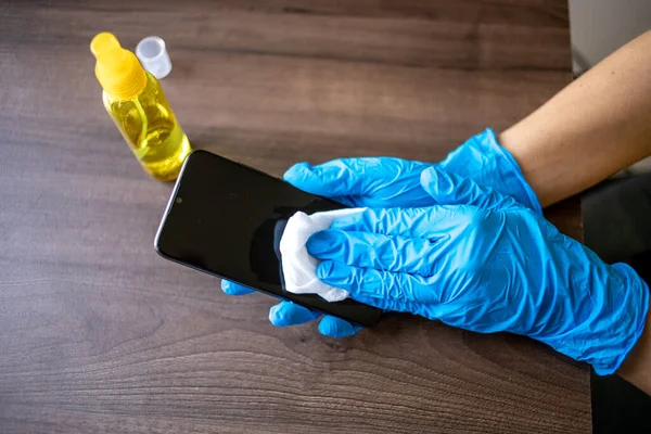 Woman's hand in blue gloves sanitizing cleaning smartphone mobile phone on wood table surface with wet wipes and alcohol