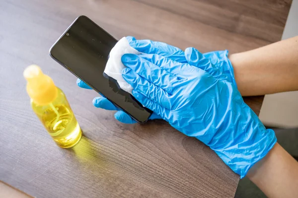 Woman's hand in blue gloves sanitizing cleaning smartphone mobile phone on wood table surface with wet wipes and alcohol