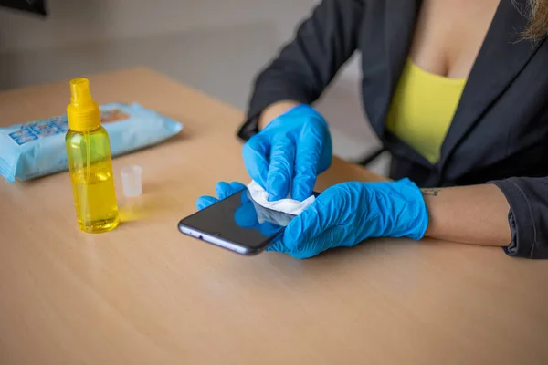 Woman\'s hand in blue gloves sanitizing cleaning smartphone mobile phone on wood table surface with wet wipes and alcohol