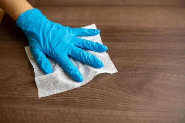 Woman's hand in blue gloves sanitizing cleaning home office wood table surface with wet wipes