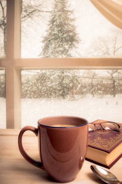 Cozy winter still-life with mug of coffee, book and reading glasses in front of a snowy window scene
