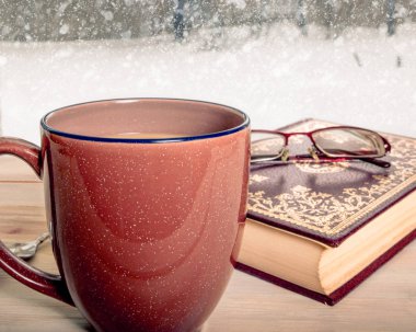 Cozy winter still-life with mug of coffee, book and reading glasses in front of a snowy window scene