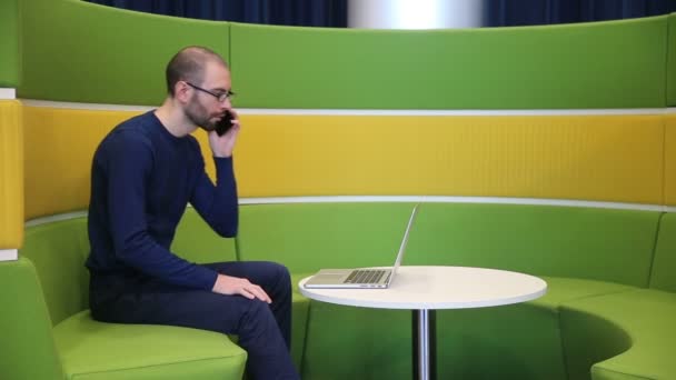 Homem careca com óculos trabalhando no laptop com telefone — Vídeo de Stock