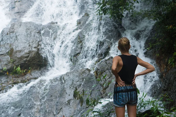 Mujer rubia de pie sobre una roca y mirando hacia arriba cerca de la cascada — Foto de Stock