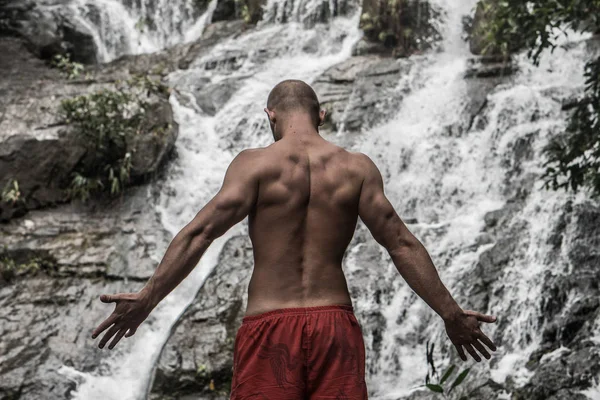 Back view of muscular man standing with raised arms near waterfall — Stock Photo, Image