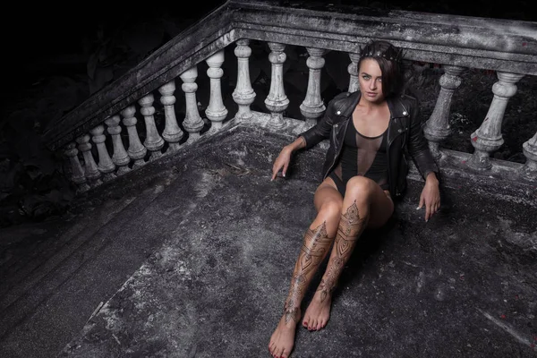 Mysterious beautiful woman in black bodysuit, leather jacket and crown with henna tattoo on her legs sitting leaning against old stone steps with ornate carved balustrade while looking at the camera — Stock Photo, Image
