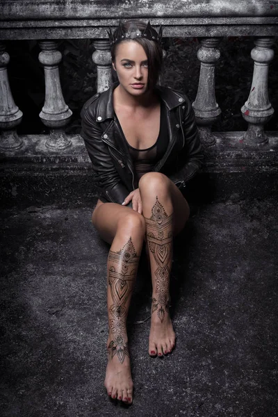 Szép nő, fekete Body, a bőrkabát és a korona henna tetoválás a lábát ülő támaszkodva régi kő lépéseket a díszes faragott korlátok, miközben nézi a kamerát — Stock Fotó