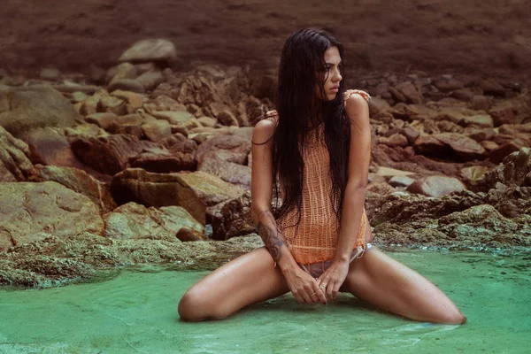 Sensuale bellezza bruna indossa top a rete arancione seduto in acqua turchese tra le rocce — Foto Stock
