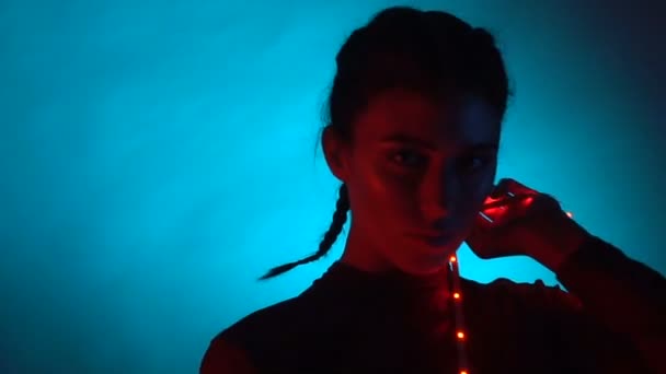 Mooie jonge sexy vrouw model spelen met rode fairy lichten, zittend op de kruk en poseren in studio in het licht van de flitser flitsen over blauwe gloeiende achtergrond — Stockvideo