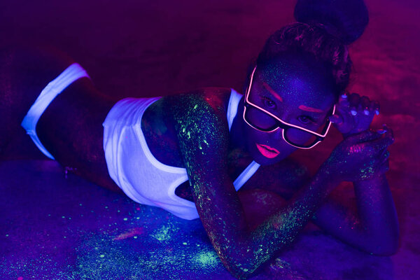 Сексуальная женщина с флуоресцентным макияжем тела и лица позирует на бетонном полу, креативный макияж отлично смотрится в ночных клубах. Вечеринка на Хэллоуин, шоу и музыка
