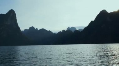 Tropikal bir göl, dağlar, uçurumlar ve kayalar su yüzeyine günbatımı arka plan, Khao Sok Milli Parkı, Surat Thani, Tayland - video yavaş tekne gezisine