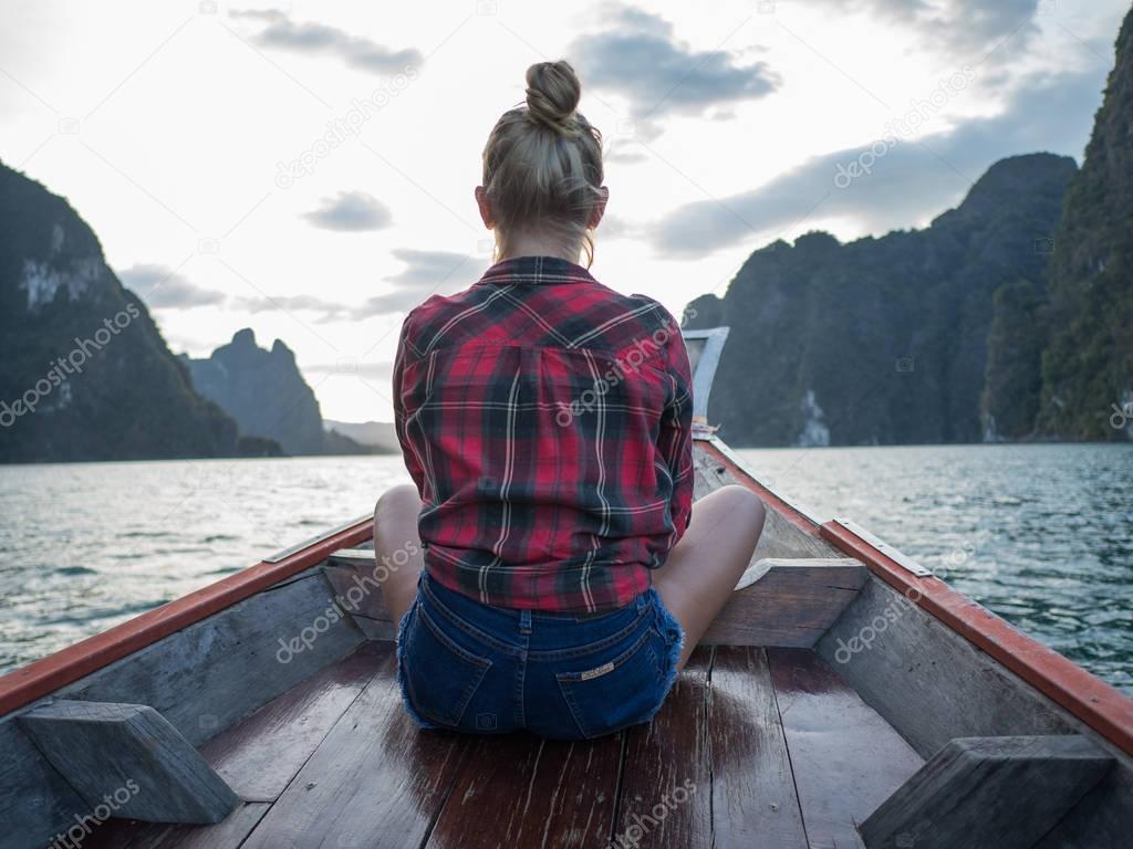 Back view of woman sitting in wood long tail boat over tropical limestone cliffs and sunset background