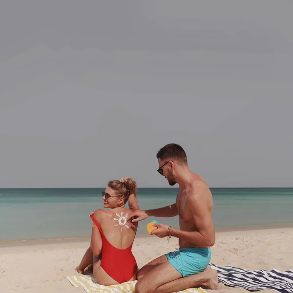 Happy couple applying sun tanning lotion on the beach, man putting sunscreen suntan cream on woman