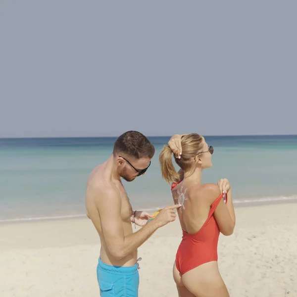 Happy couple applying sun tanning lotion on the beach, man putting sunscreen suntan cream on woman