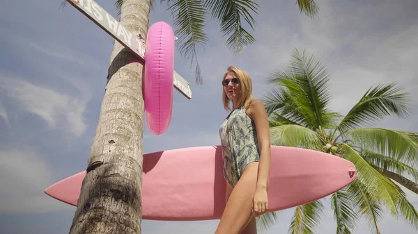 水着とサングラス ピンク サーフボードとの日当たりの良い夏の日の間に ヤシの木と空の背景歩いて幸せな若い女 — ストック写真