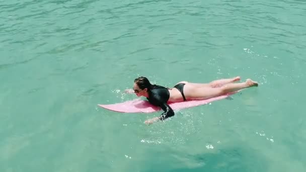 水着とサングラスに驚くほど澄んだ海水 スローモーションのビデオで彼女の休日を楽しんでいるピンクのサーフボードの美しい若い女性の空中のトップ ビュー — ストック動画